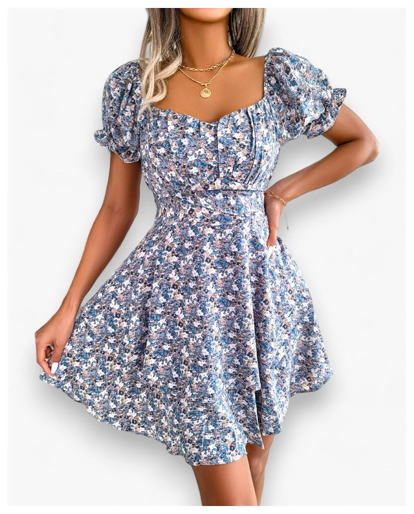 A woman displays a charming blue Floral Mini Dress with High Waist and Swing Style from Eternal Gleams. The dress, featuring puffed short sleeves, a sweetheart neckline, and a fitted bodice with a flared skirt, offers a cool and calming style. The blue tone provides a serene and sophisticated look, making it perfect for any occasion. Paired with a gold necklace, the dress’s elegant and graceful nature shines through.