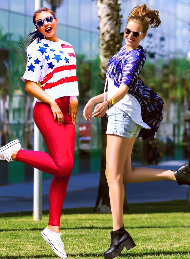 Two stylish women in casual outfits joyfully jumping outdoors. The first woman, on the left, wears sunglasses, a white crop top with blue stars and red stripes, red leggings, and white sneakers. The second woman, on the right, sports sunglasses, an updo hairstyle, a blue and white patterned top, denim shorts, and black ankle boots. They are set against a backdrop of palm trees and modern glass buildings, enjoying a sunny day.